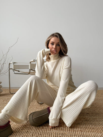 Woman wearing a white ribbed sweater and pants sitting on a textured rug.