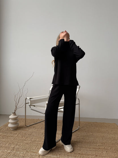 Person wearing a black outfit in a minimalistic room with a chair and vase.