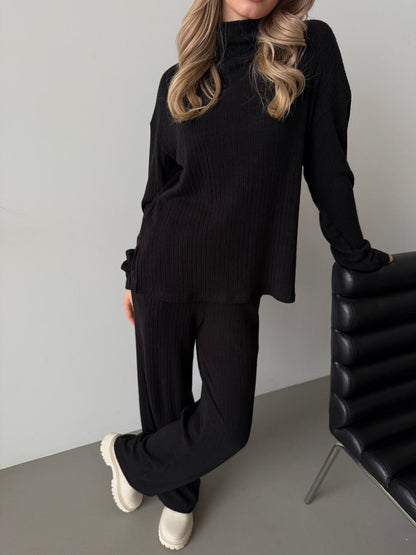 Person wearing a black ribbed sweater and pants with white shoes, standing against a gray wall.
