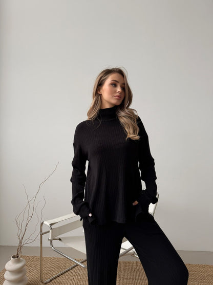 Woman wearing a black turtleneck sweater and pants in a minimalistic setting.