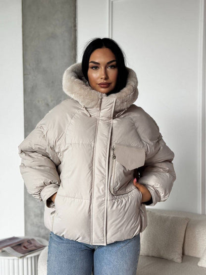 Woman wearing a beige puffer jacket with a fur-lined hood in a room.