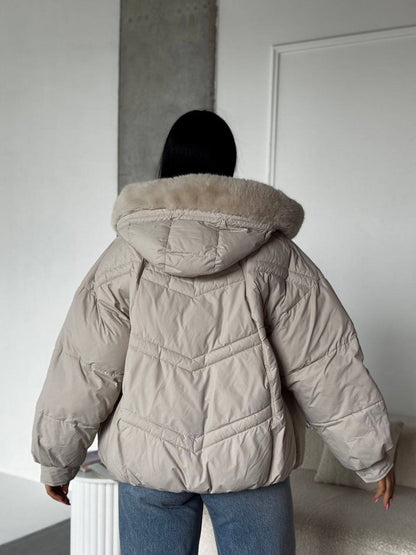 Person wearing a beige puffer jacket with a fur-lined hood in a minimal indoor setting.