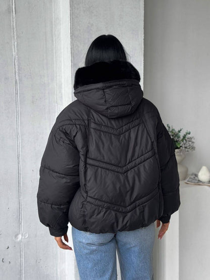 Person wearing a black puffer jacket with a fur-lined hood against a light gray wall.