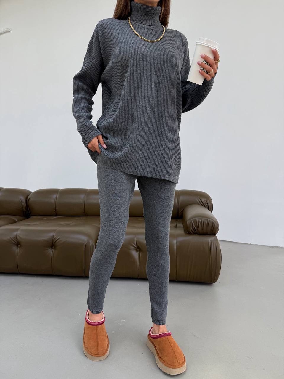 Person wearing a gray sweater and leggings set with brown shoes, holding a white cup indoors.