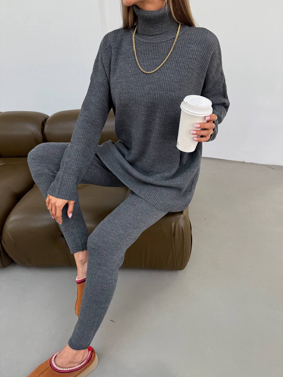 Person wearing a gray knit outfit sitting on a brown couch holding a white coffee cup.
