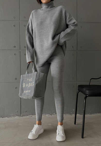 Person wearing a gray outfit holding a bag with text against a gray wall.