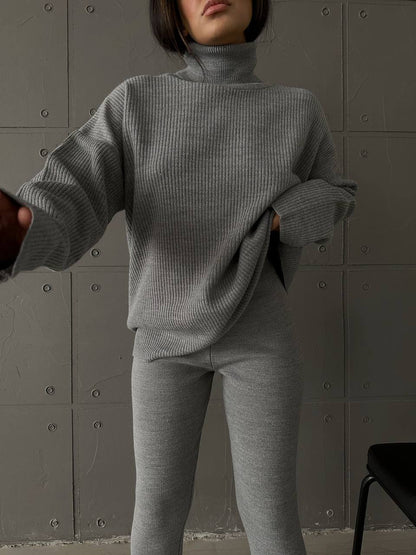 Person wearing a gray ribbed sweater and pants against a textured wall.