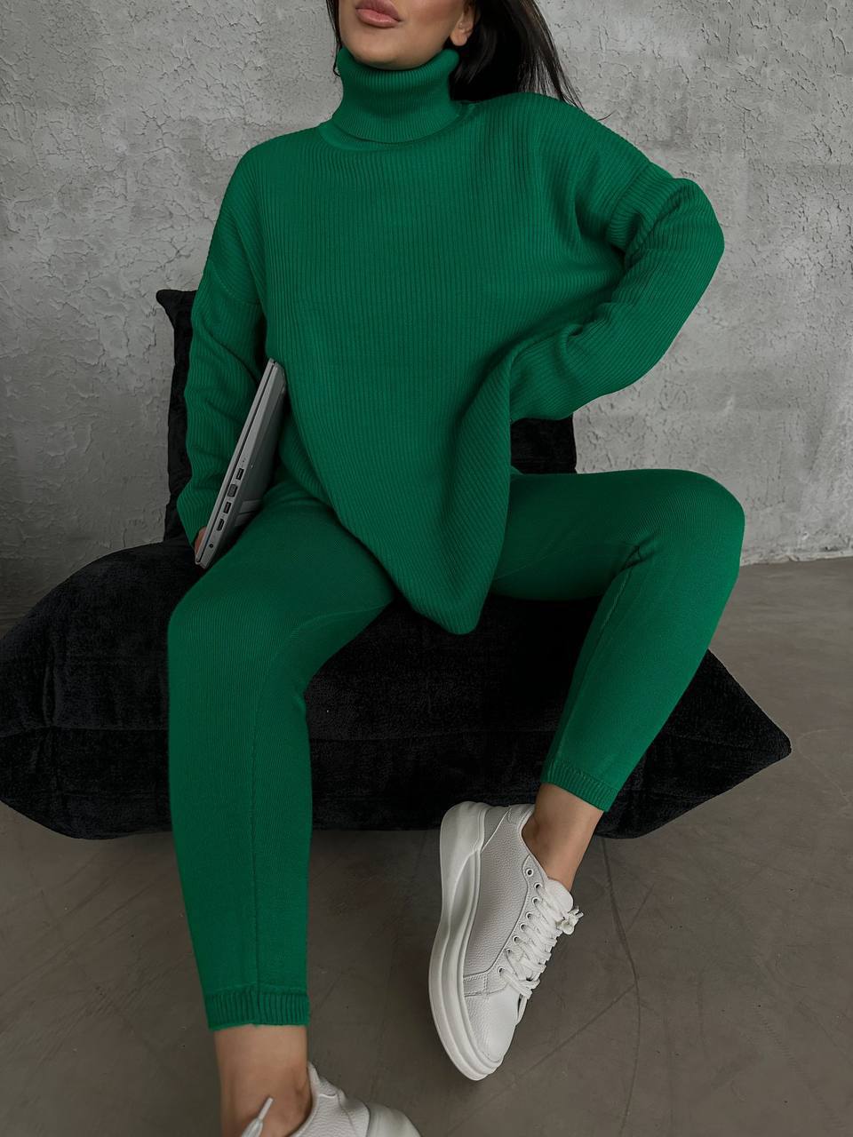 Person wearing a green outfit with white sneakers sitting on a black chair against a gray wall.