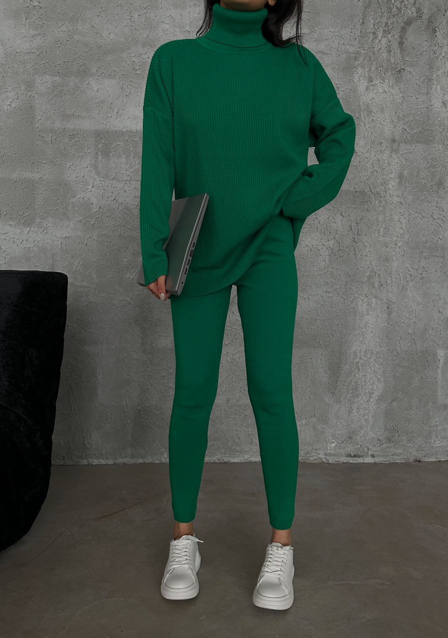 Person wearing a green outfit holding a laptop against a gray concrete wall.