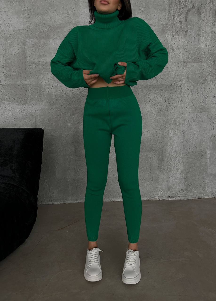 Person wearing a green outfit with a turtleneck sweater and leggings against a gray wall.