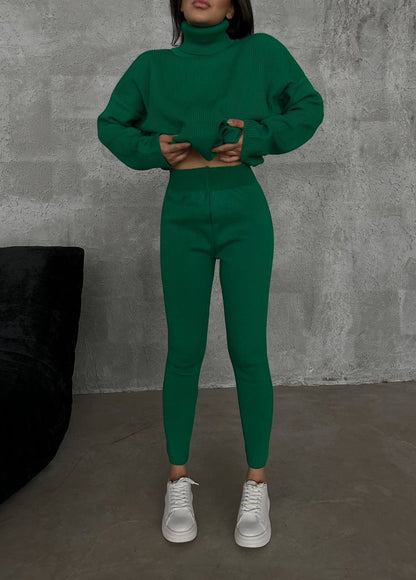 Person wearing a green outfit with a turtleneck sweater and leggings against a gray wall.