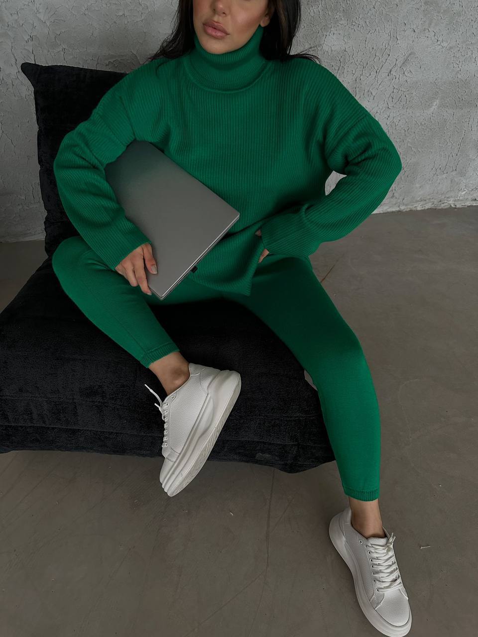 Person wearing a green outfit sitting on a black cushion with a gray laptop.