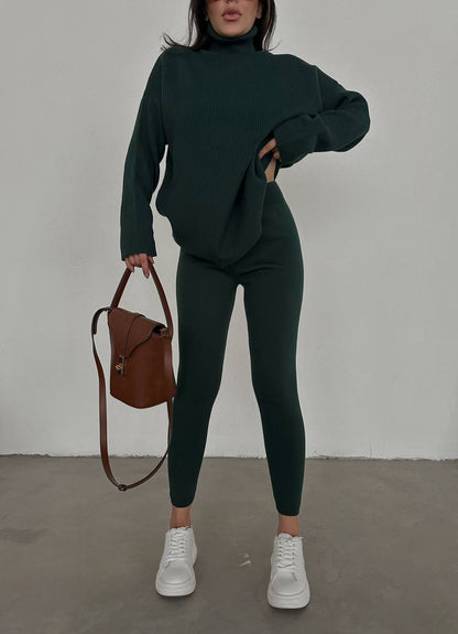 Person wearing a dark green outfit with a brown bag against a plain background