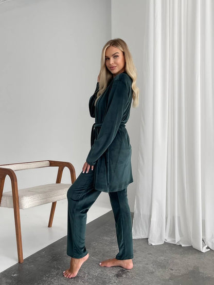 Woman in a dark green outfit standing in a room with a white curtain and wooden chair.
