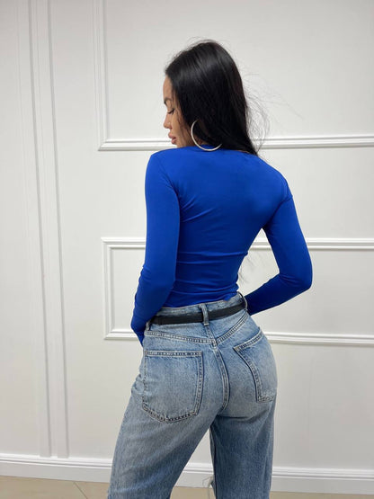 Person wearing a blue long-sleeve top and light blue jeans against a white paneled wall.