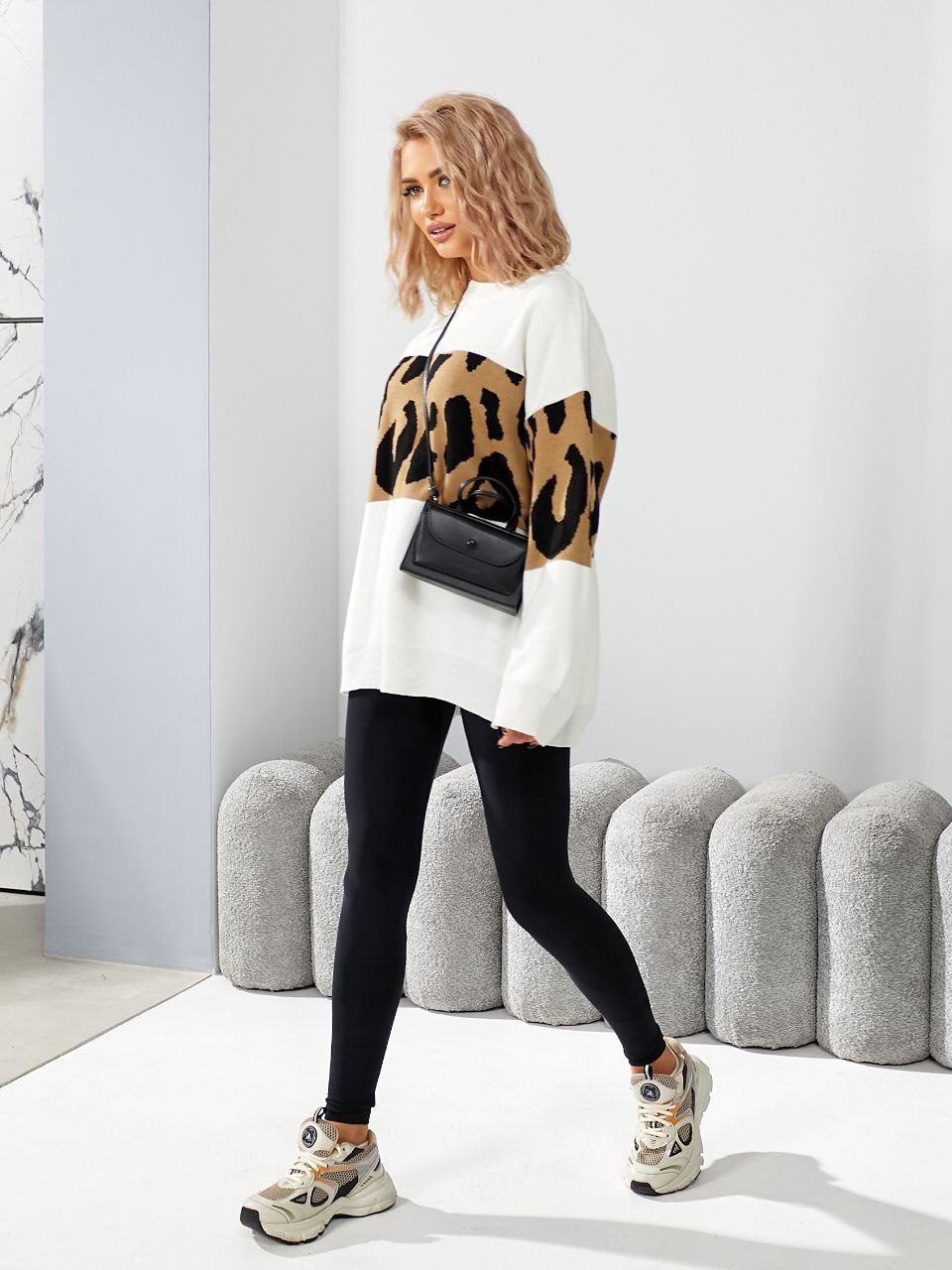 Woman wearing a white sweater with leopard print accents, black leggings, and sneakers in a modern room.