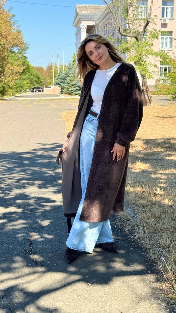 Woman wearing a long brown coat standing outdoors with trees and buildings in the background
