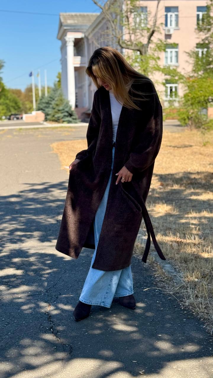 Person wearing a long dark coat in an outdoor setting with buildings and trees in the background.