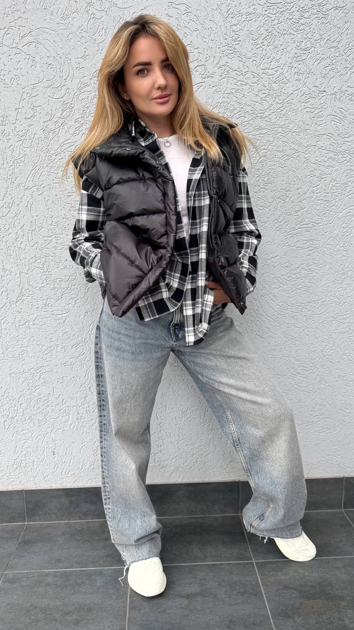 Woman wearing a plaid shirt, black vest, and gray pants against a gray wall.