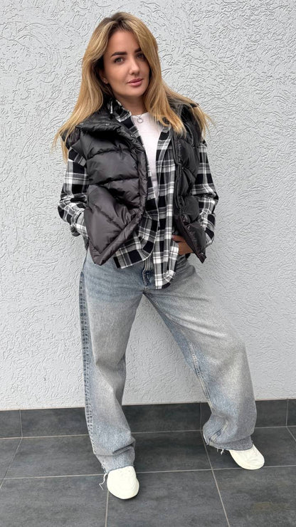 Woman wearing a plaid shirt, black vest, and gray pants against a gray wall.
