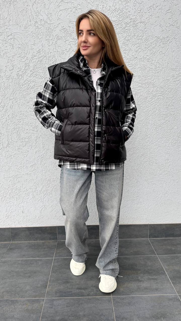 Woman wearing a black puffer vest over a plaid shirt with gray pants against a gray wall.