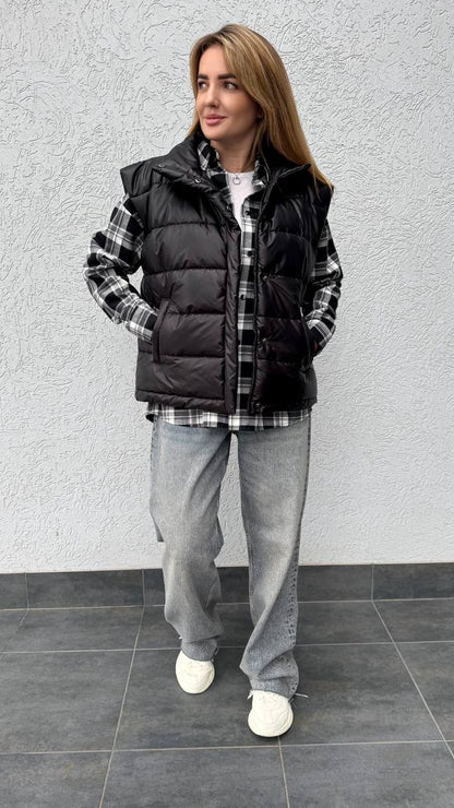 Woman wearing a black puffer vest over a plaid shirt with gray pants against a gray wall.