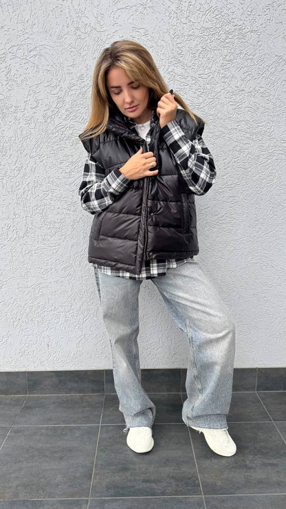 Woman wearing a black puffer vest over a plaid shirt with gray pants against a gray wall.