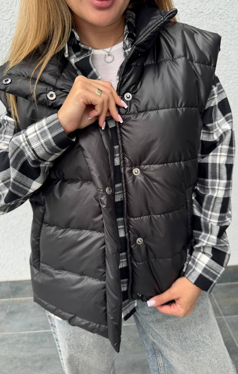 Person wearing a black puffer vest over a plaid shirt with a neutral background
