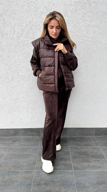 Woman wearing a brown puffer jacket and pants against a gray wall.
