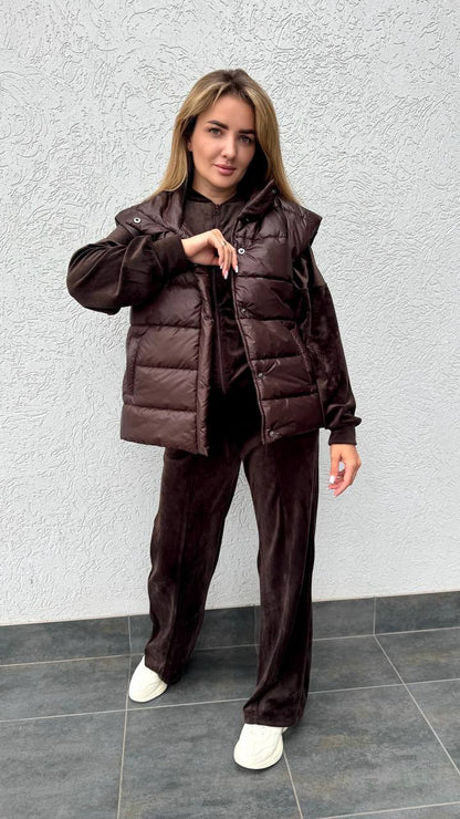 Woman wearing a brown puffer jacket and pants against a gray wall.