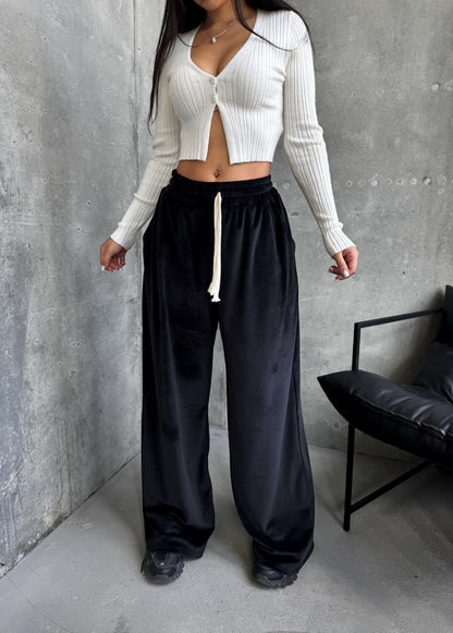 Person wearing a white ribbed cardigan and black pants against a gray wall.