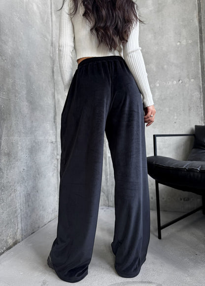 Person wearing black wide-leg pants and a light-colored sweater against a gray wall.
