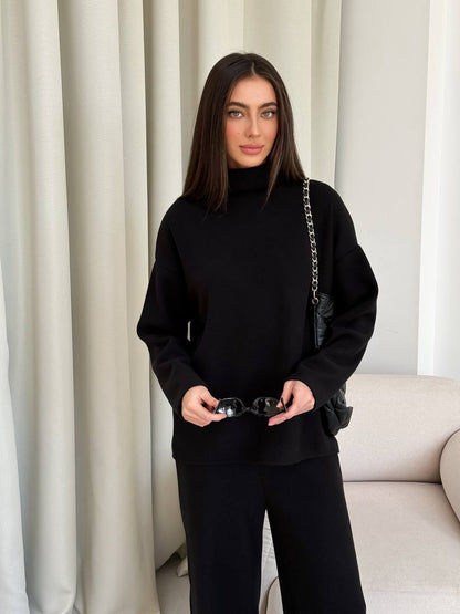 Woman in a black outfit holding sunglasses in front of a neutral curtain.