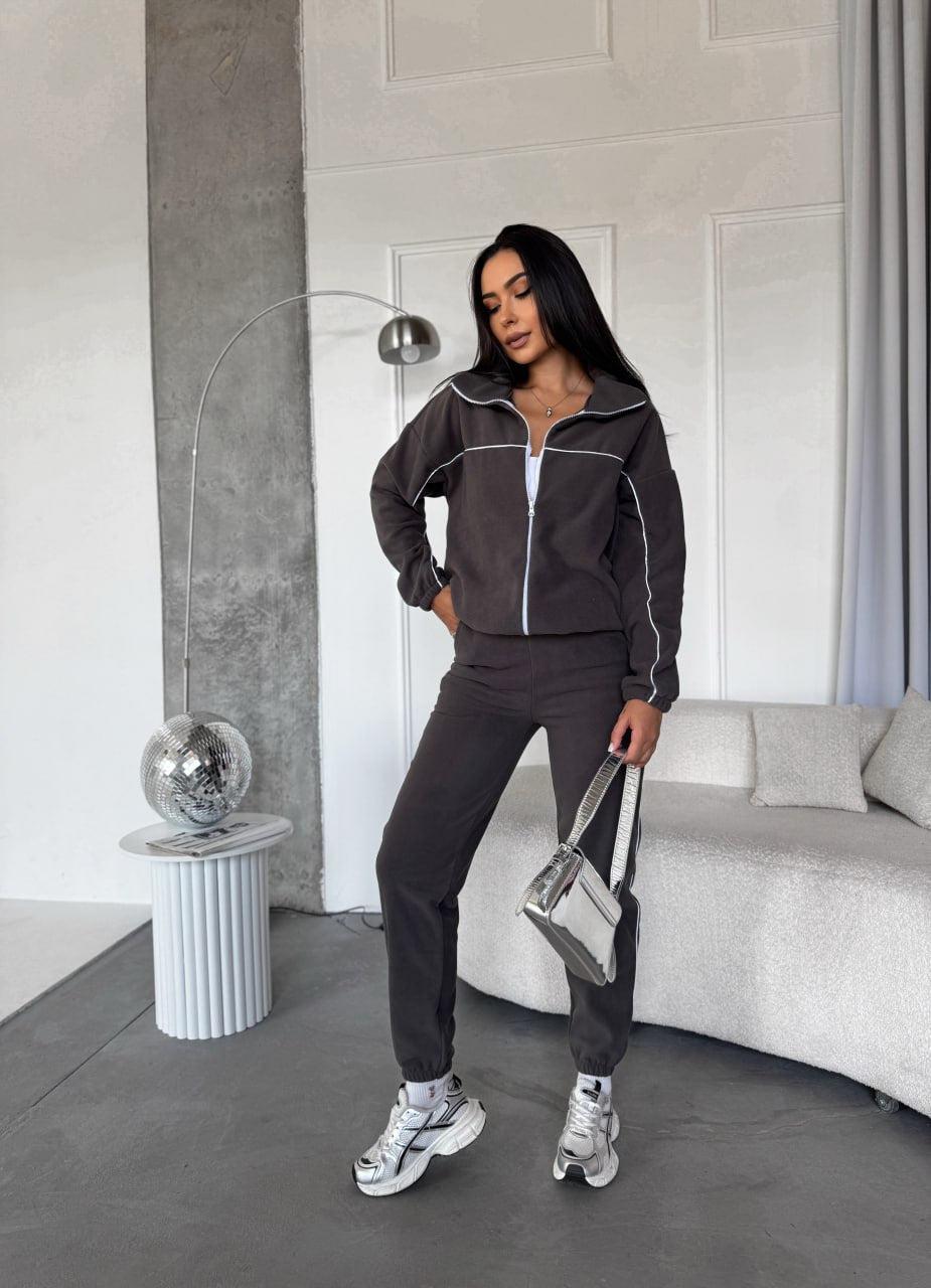 Woman in a dark gray tracksuit standing in a modern room with a sofa and decorative items.