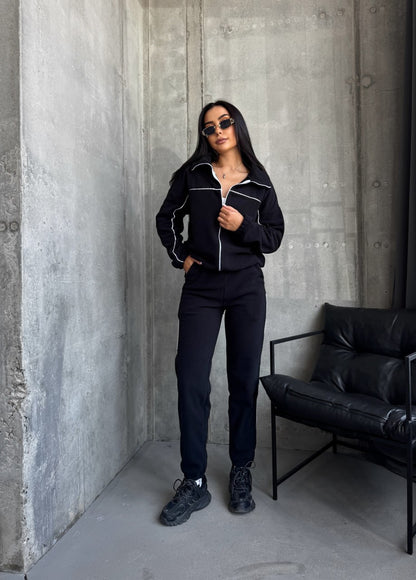 Person wearing a black tracksuit standing against a concrete wall with a black chair.