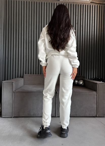Person wearing a white tracksuit standing in a room with a gray couch and striped wall.