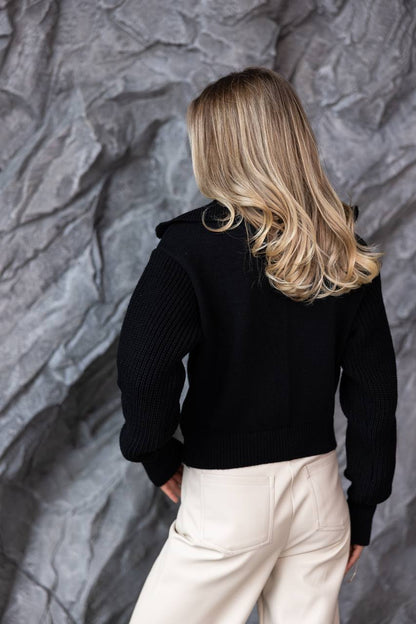 Person wearing a black jacket and white pants against a textured gray wall