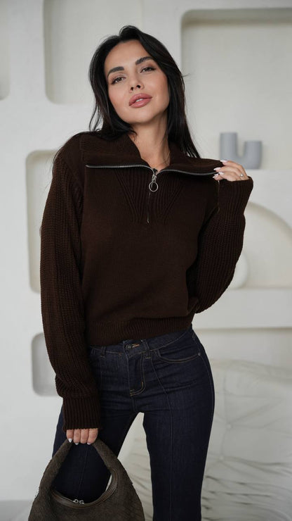 Woman wearing a brown zip-up sweater and dark jeans against a white textured wall.