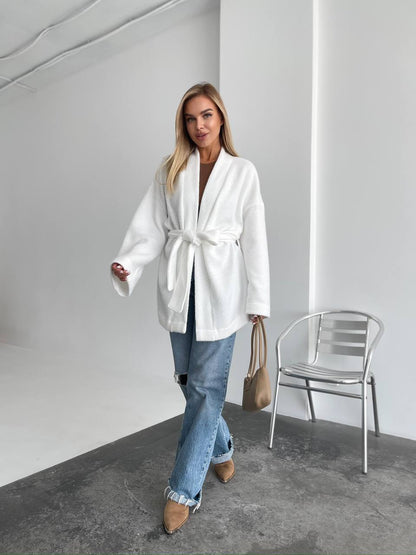 Woman wearing a white robe in a minimalistic room with a chair and bag.