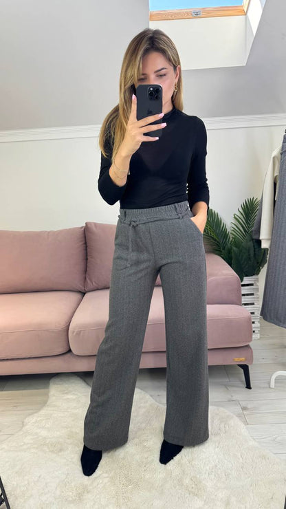 Woman taking a mirror selfie wearing a black top and gray pants in a room with a pink couch and white walls.