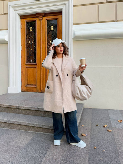 Woman in a beige coat holding a coffee cup and a bag, standing in front of a building entrance.