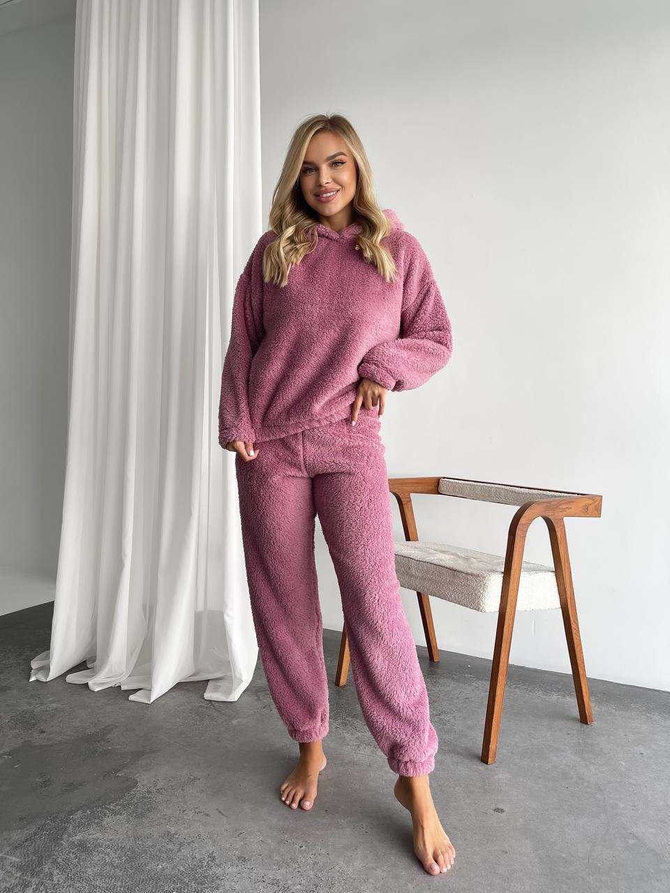 Woman wearing a pink fleece loungewear set standing in a room with white curtains and a wooden chair.