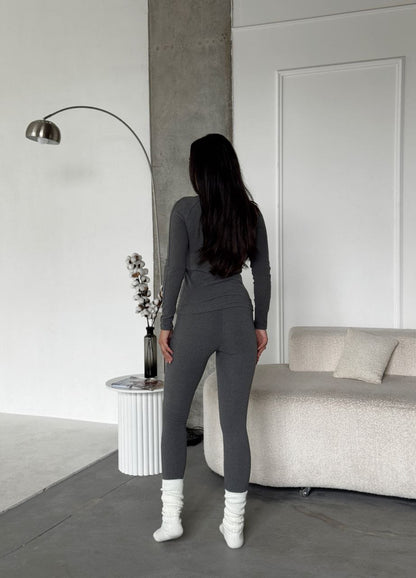 Person wearing a gray outfit standing in a minimalistic room with a sofa and lamp.