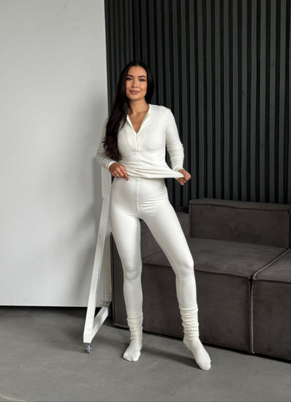 Woman in a white outfit standing in a modern room with a gray sofa and black vertical blinds.