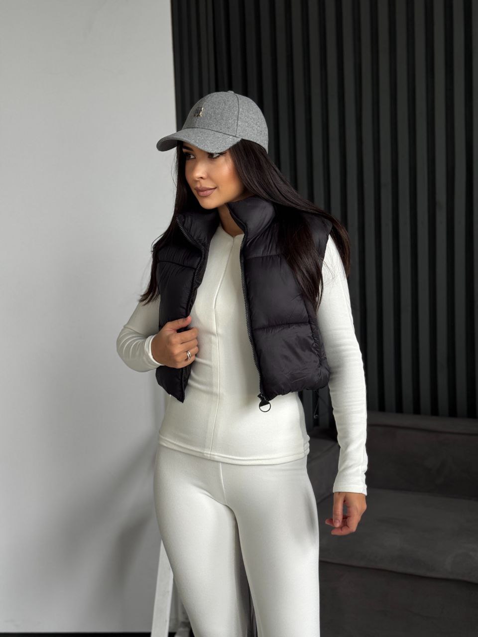 Person wearing a black puffer vest over a white outfit with a gray cap indoors.