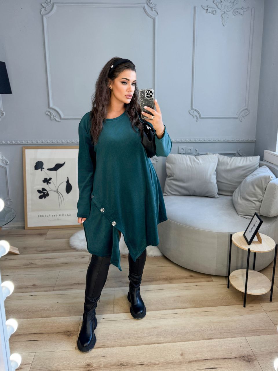Woman taking a mirror selfie wearing a green dress and black boots in a stylish room.