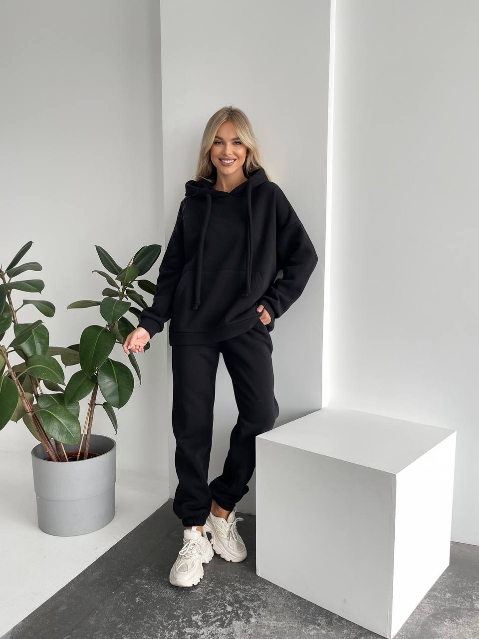 Person wearing a black hoodie and pants standing next to a plant in a minimalistic setting