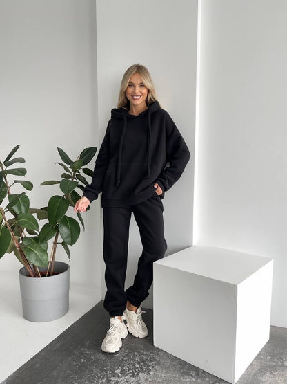 Person wearing a black hoodie and pants standing next to a plant in a minimalistic setting