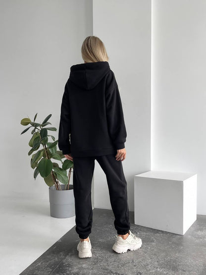 Person wearing a black hoodie and pants standing in a minimalistic room with a plant and white cube.
