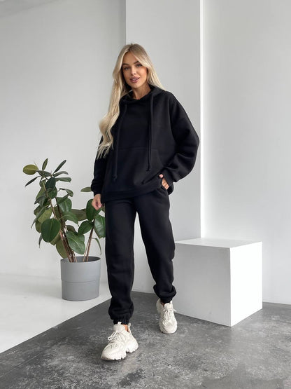 Person wearing a black hoodie and pants in a minimalistic setting with a plant.